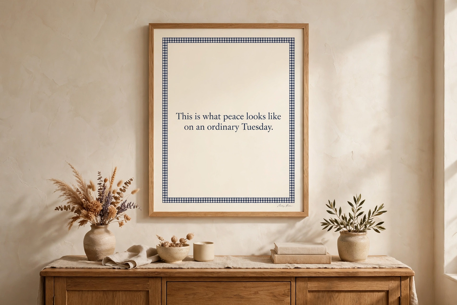 A framed print with a navy gingham border reading 'This is what peace looks like on an ordinary Tuesday.' hanging above a wooden sideboard